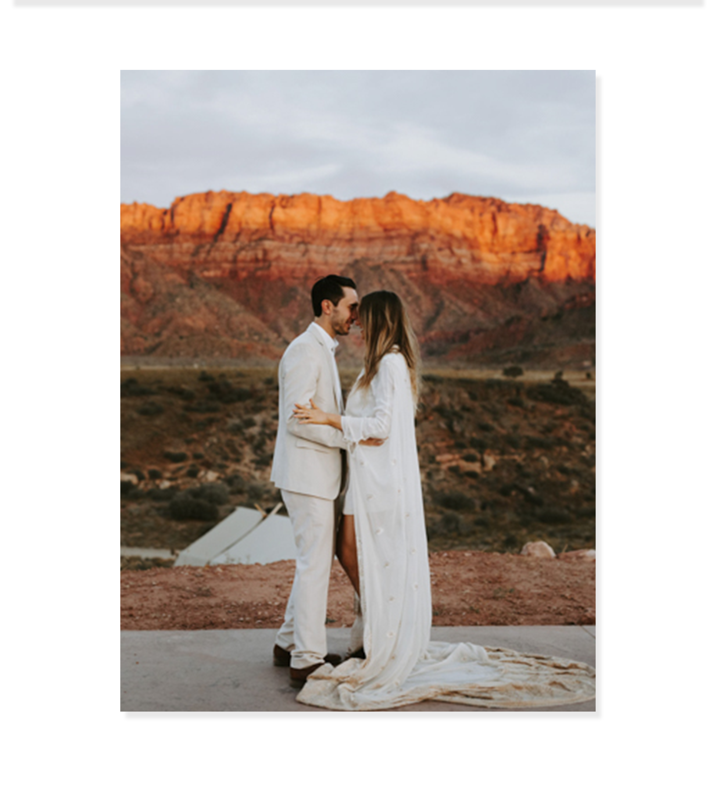Un cuplu îmbrăcat în alb stă aproape unul de altul în aer liber, cu stânci roșii și un cer înnorat în spatele lor - un moment uimitor, perfect pentru o culoare pe pânză sau pentru adăugarea la colecția dvs. de artă.